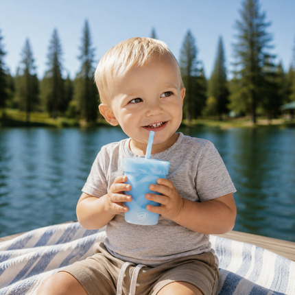 A young toddler with blond hair sits on a striped blanket by a lake, smiling and holding a light blue straw cup with a blue drink. Trees and blue water are visible in the background.