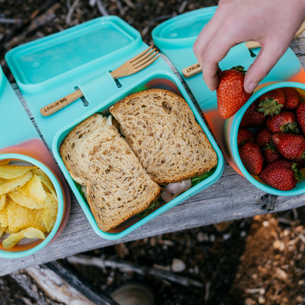 A turquoise silicone lunch box set arranged on a weathered wooden picnic blanket outdoors. The set includes a square lunch box with two slices of bread, a round bowl of potato chips, a round bowl of strawberries being held by a hand, and another lunch box lid in the background.