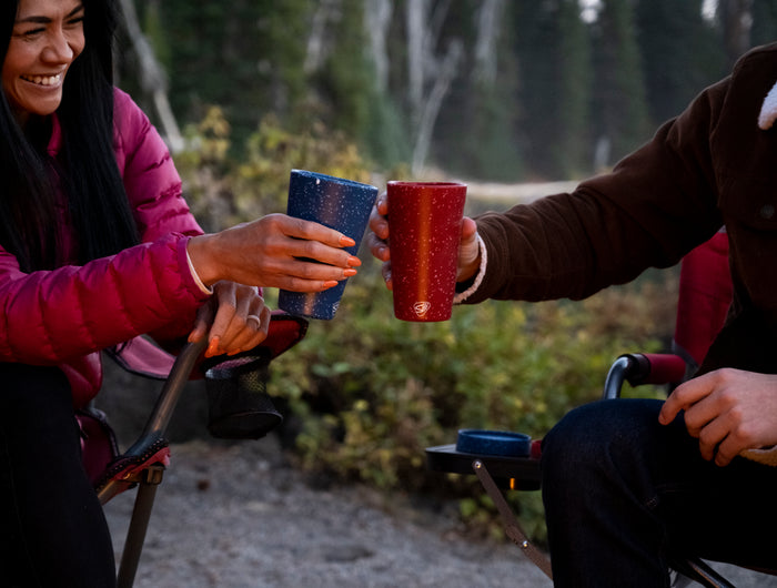 Two people sitting outdoors, clinking mugs together in a forest setting.