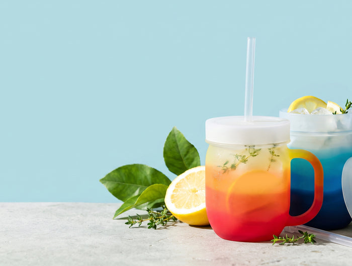 Two colorful layered drink mugs with straws on a light stone surface against a pale blue background. The front mug is red, orange, and yellow, and the rear mug is blue and white with lemon slices and green leaves around it.