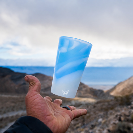 A hand holding a frosted silicone tumbler with a blue and white marbled swirl pattern, set against a blurred mountain landscape under a cloudy sky.