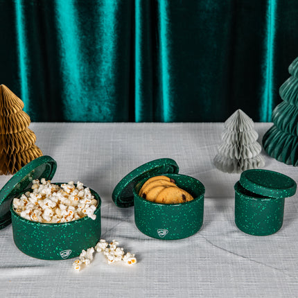 Three round green glittery snack boxes with lids, shown open with popcorn and cookies inside, alongside decorative paper holiday trees in gold, green, and silver on a light gray tablecloth in front of a dark green velvet curtain.