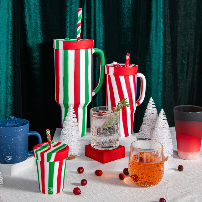 Decorative setup with Christmas-themed mugs, glasses, and small trees against a green curtain.