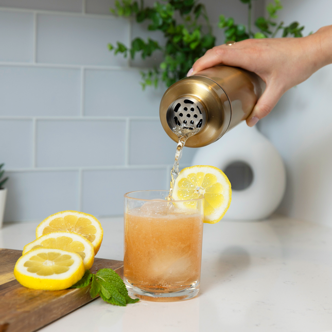 Person pouring a drink from a gold shaker into a glass with lemon slices on a kitchen counter.