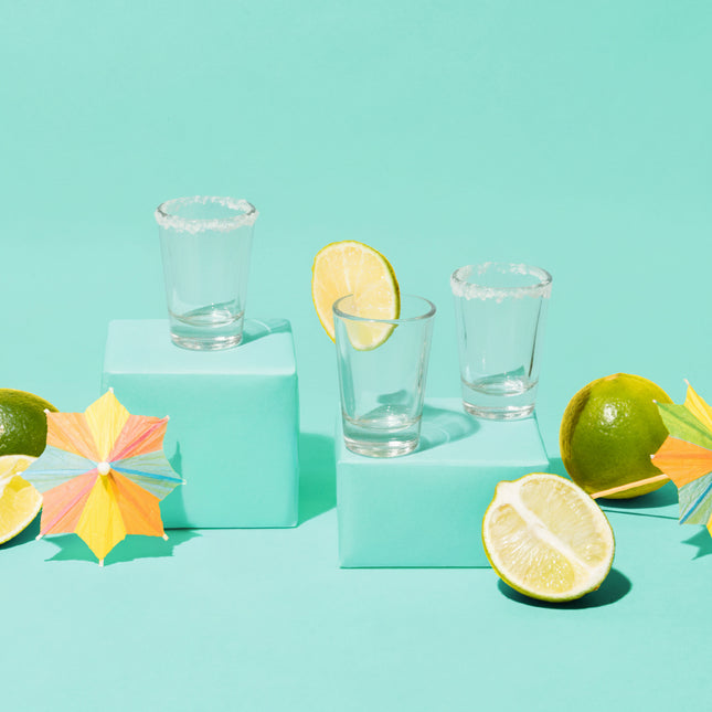 Clear shot glasses on a turquoise platform with limes and colorful leaves on a light blue background